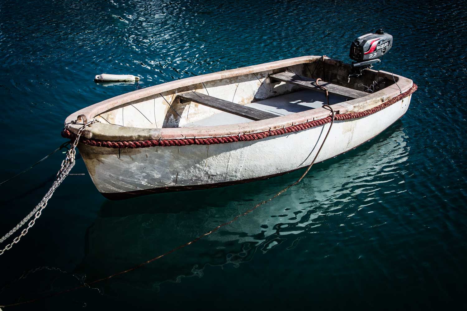 Small motorboat tied to a chain floating on clear blue water.