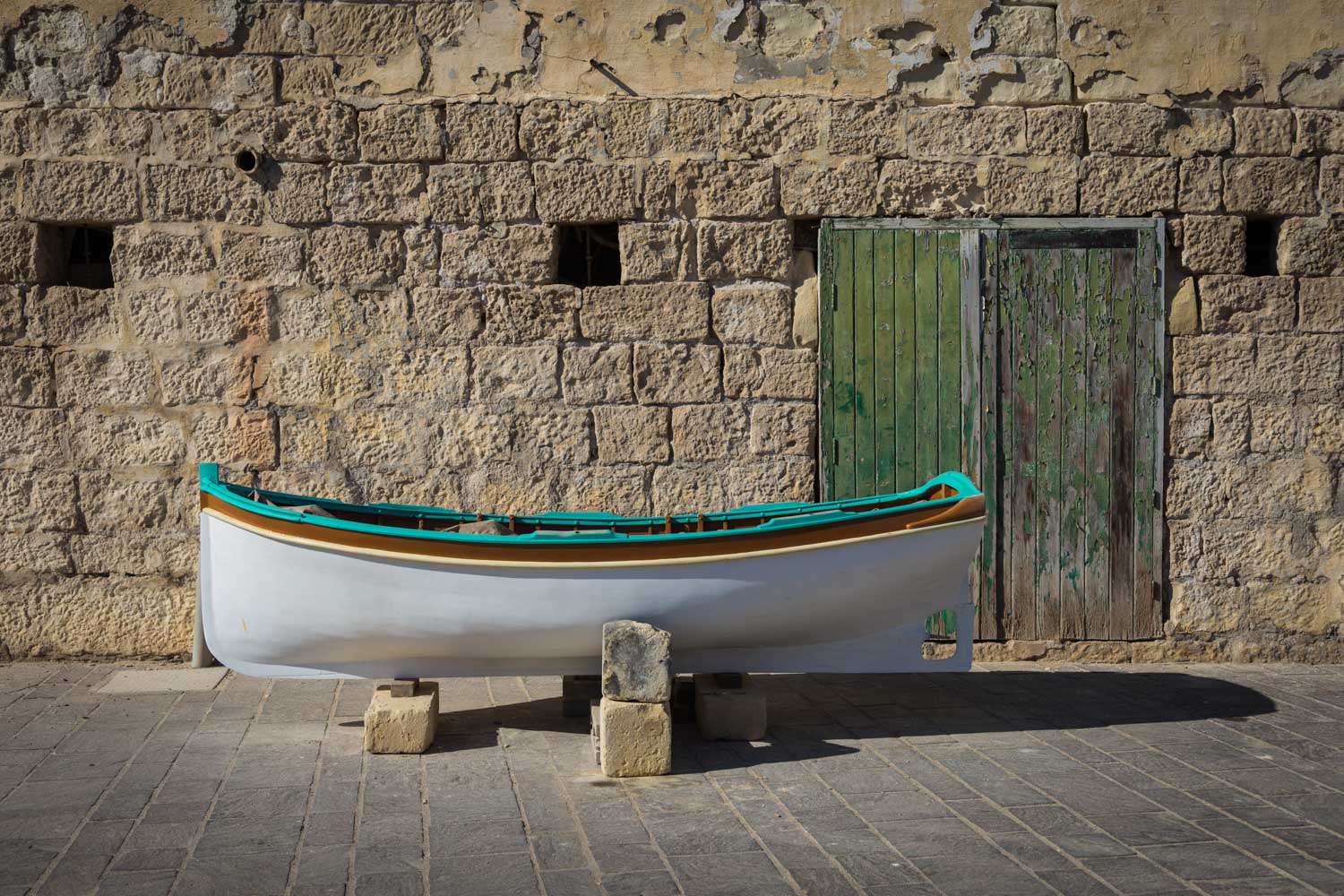 Small white and blue boat on blocks, against a rustic stone wall and weathered green doors.
