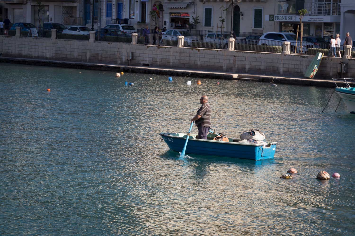 Man rowing small blue boat on calm urban waterfront, buildings and cars in background.