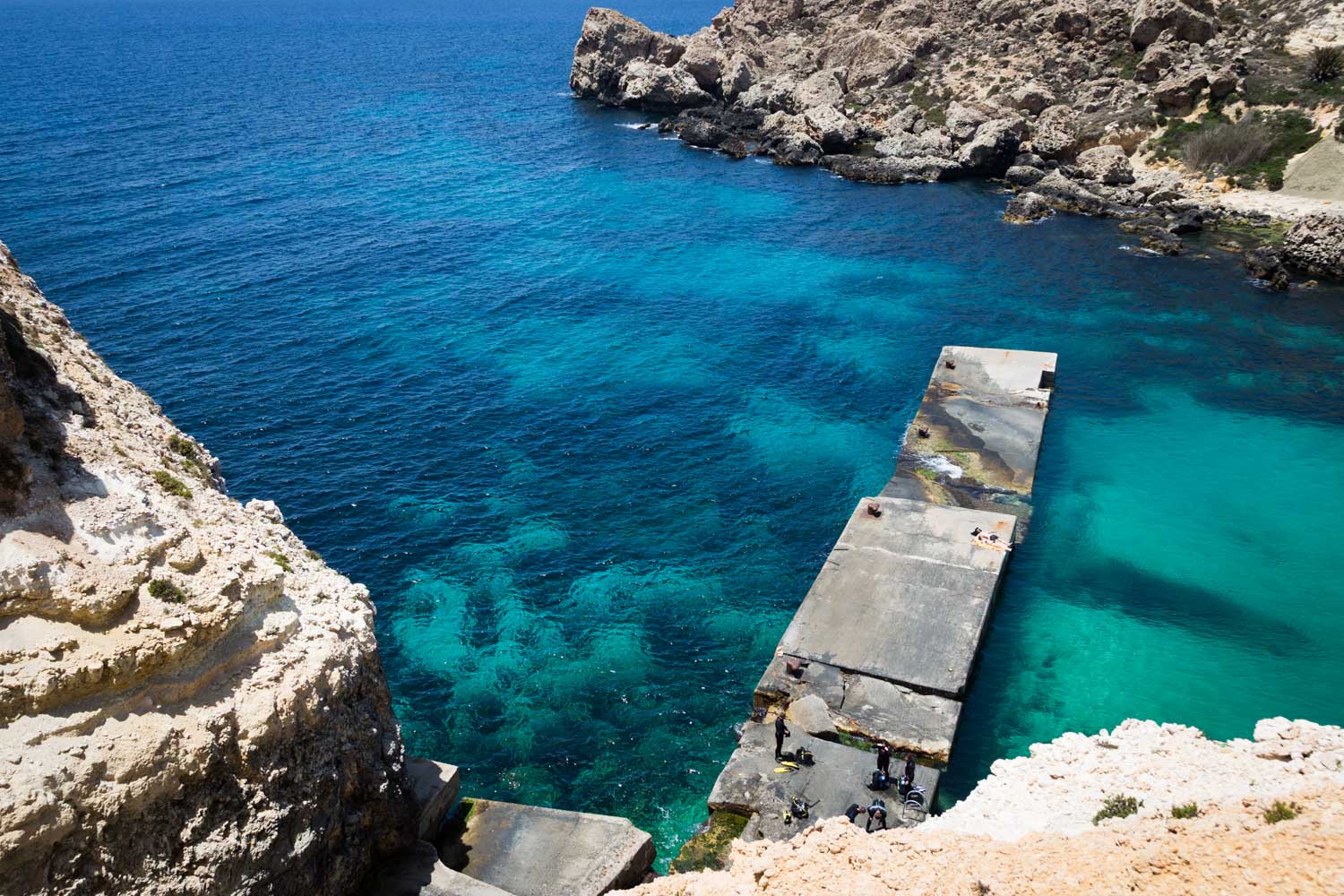 Cliffside view of a turquoise bay with rocky shoreline and a concrete pier, clear waters ideal for diving or snorkeling.