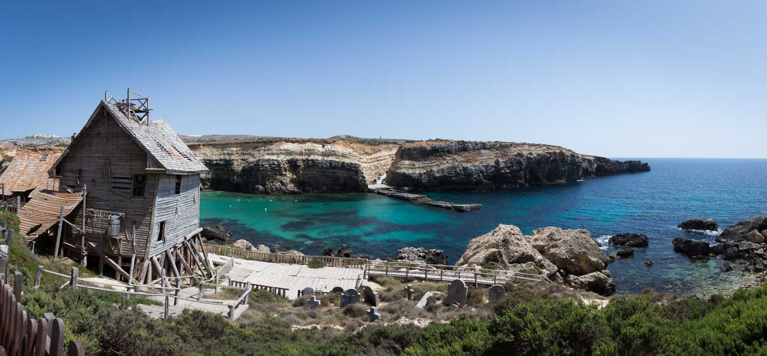 Rustic wooden house overlooking scenic turquoise bay and rocky cliffs under clear blue sky.