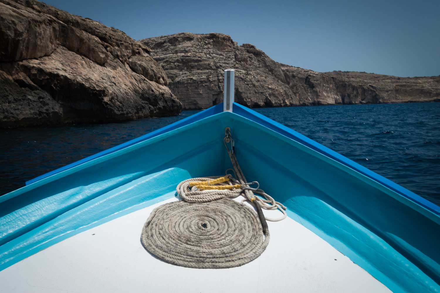 Front of a blue boat navigating through a rocky seascape under a clear sky, with coiled rope on the deck.
