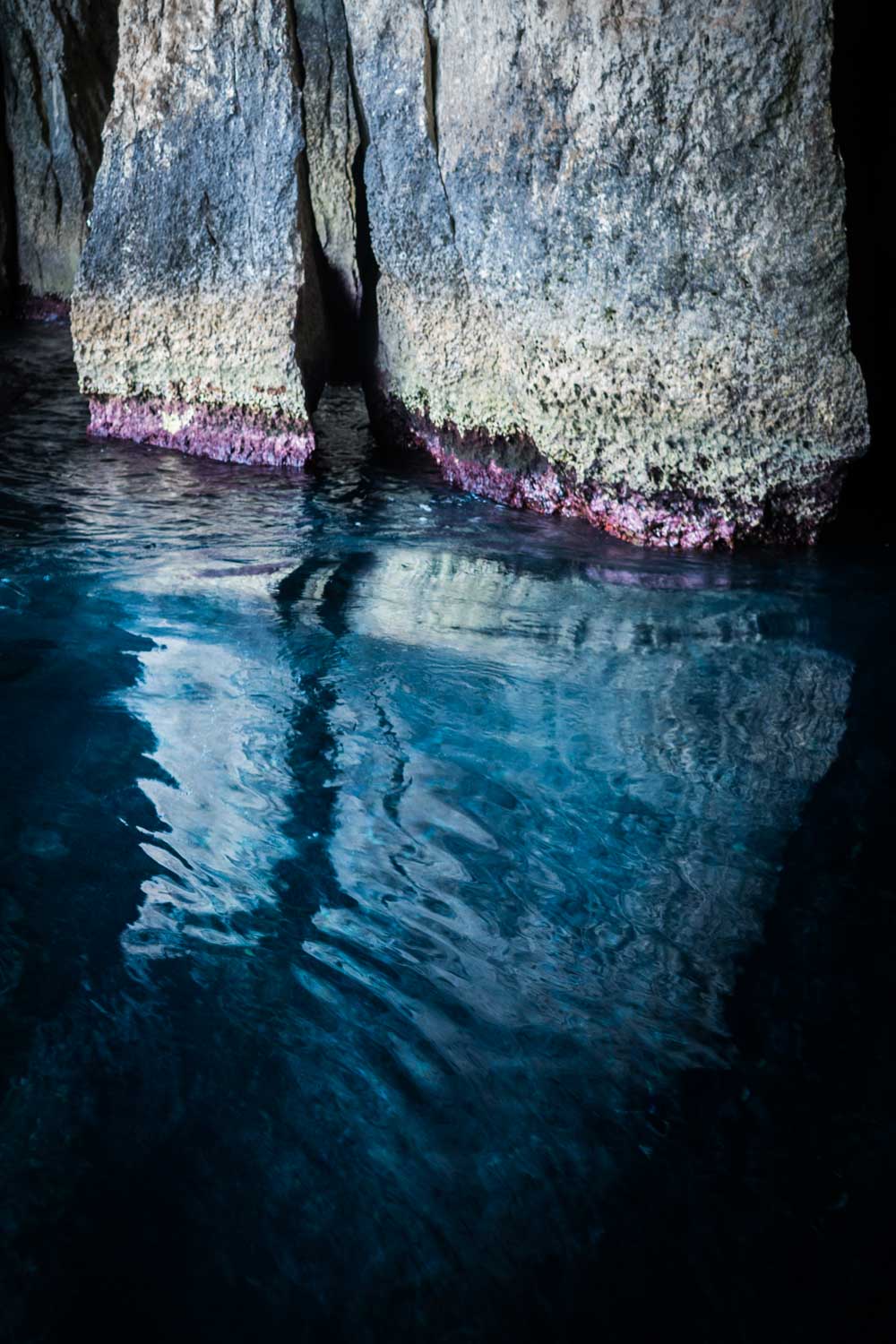 Clear blue water reflecting onto rugged cave walls with colorful algae at the base.