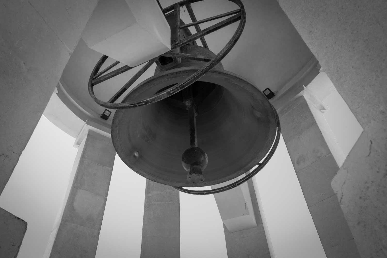 Black and white close-up of a large church bell viewed from below with stone columns in the background.