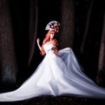 Red-haired woman in a flowing white gown and floral headpiece posing elegantly among tall trees at night.