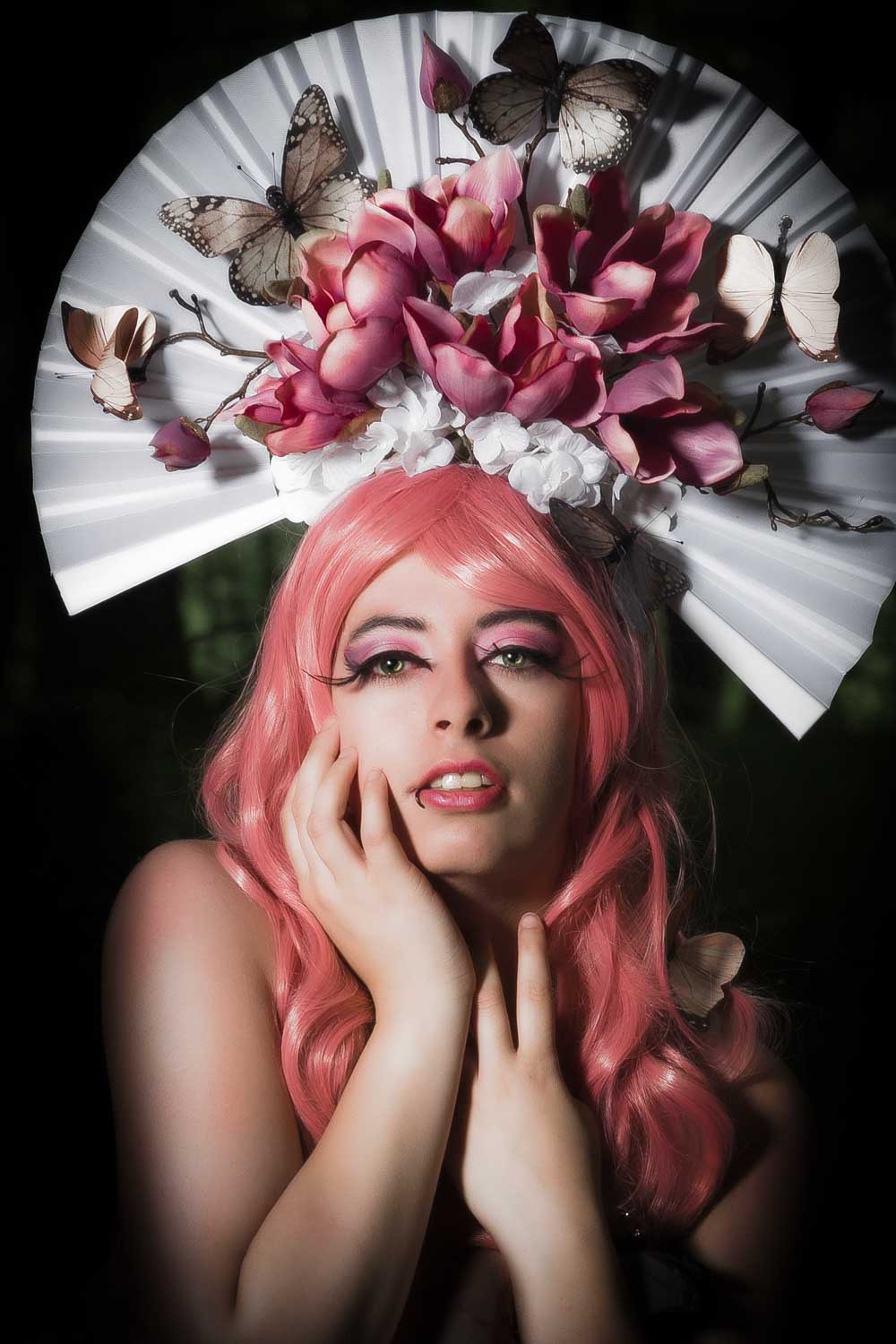 Woman in pink wig with floral and butterfly headdress, dramatic makeup, artistic portrait photography.
