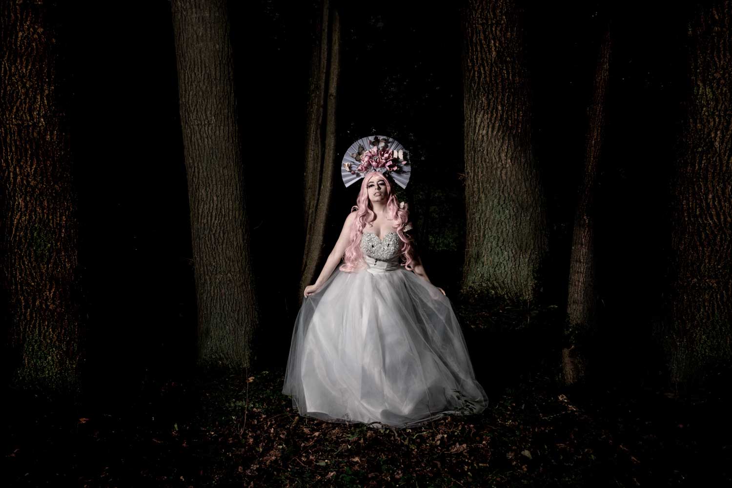 Fantasy-themed woman in a white gown and floral headdress stands in a dark forest between trees.