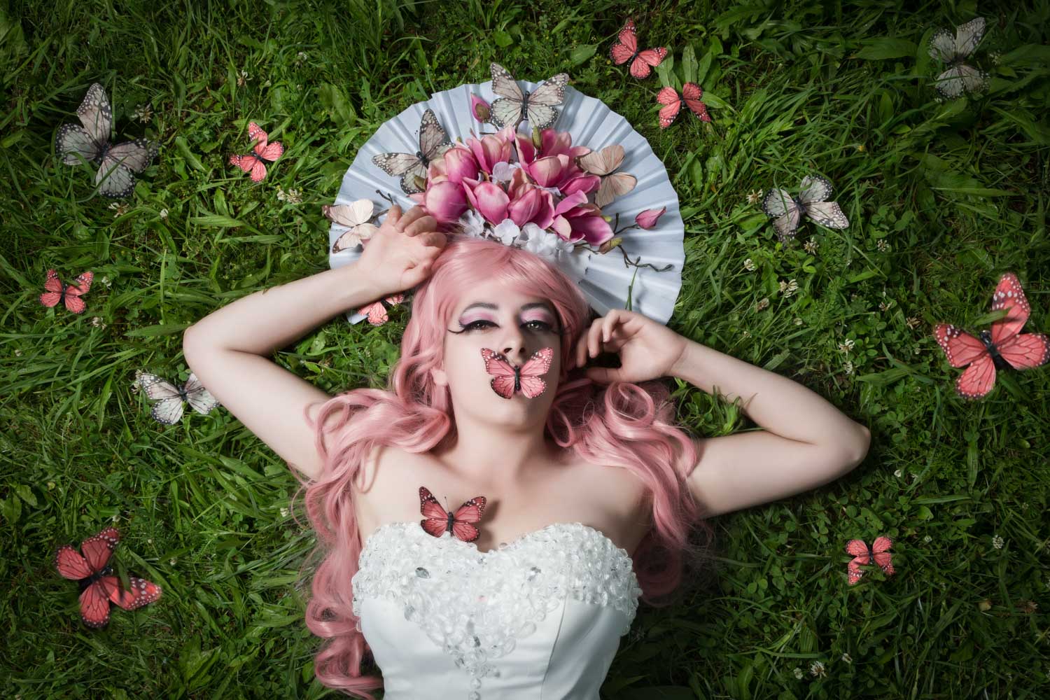 Woman with pink hair and butterfly headdress lying on grass, surrounded by colorful butterflies.
