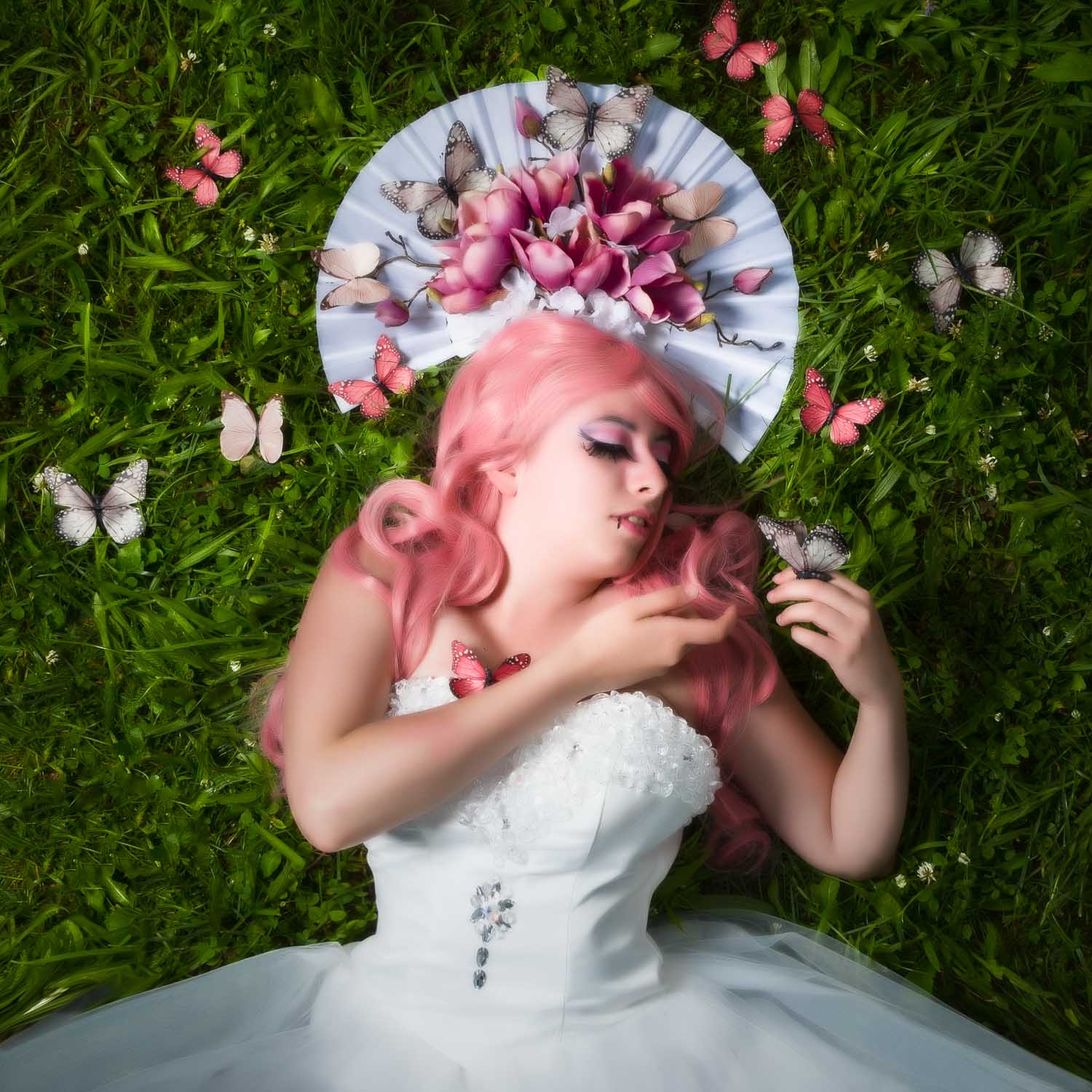 Woman in a white dress with pink hair lies on grass surrounded by butterflies and a floral fan headpiece.