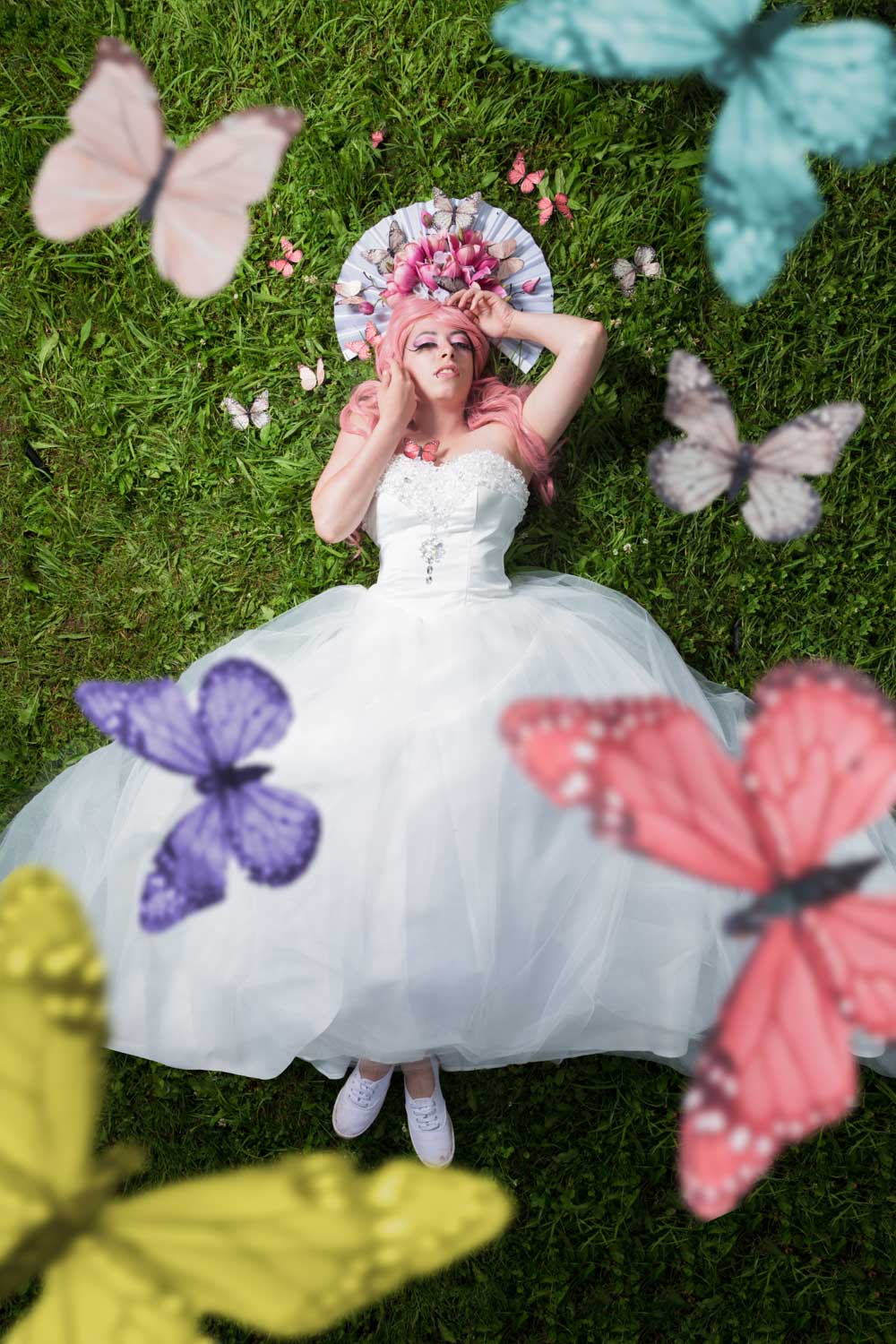 Woman in white gown with pink hair, lying on grass surrounded by colorful butterflies and holding a floral fan.