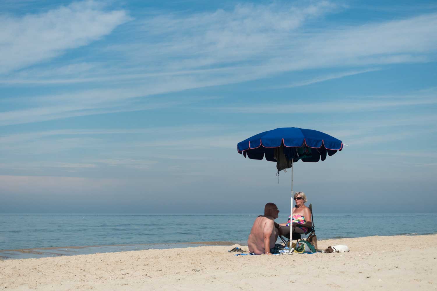 Couple relaxing under blue beach umbrella on a sandy shore with a calm ocean and cloudy sky in the background.