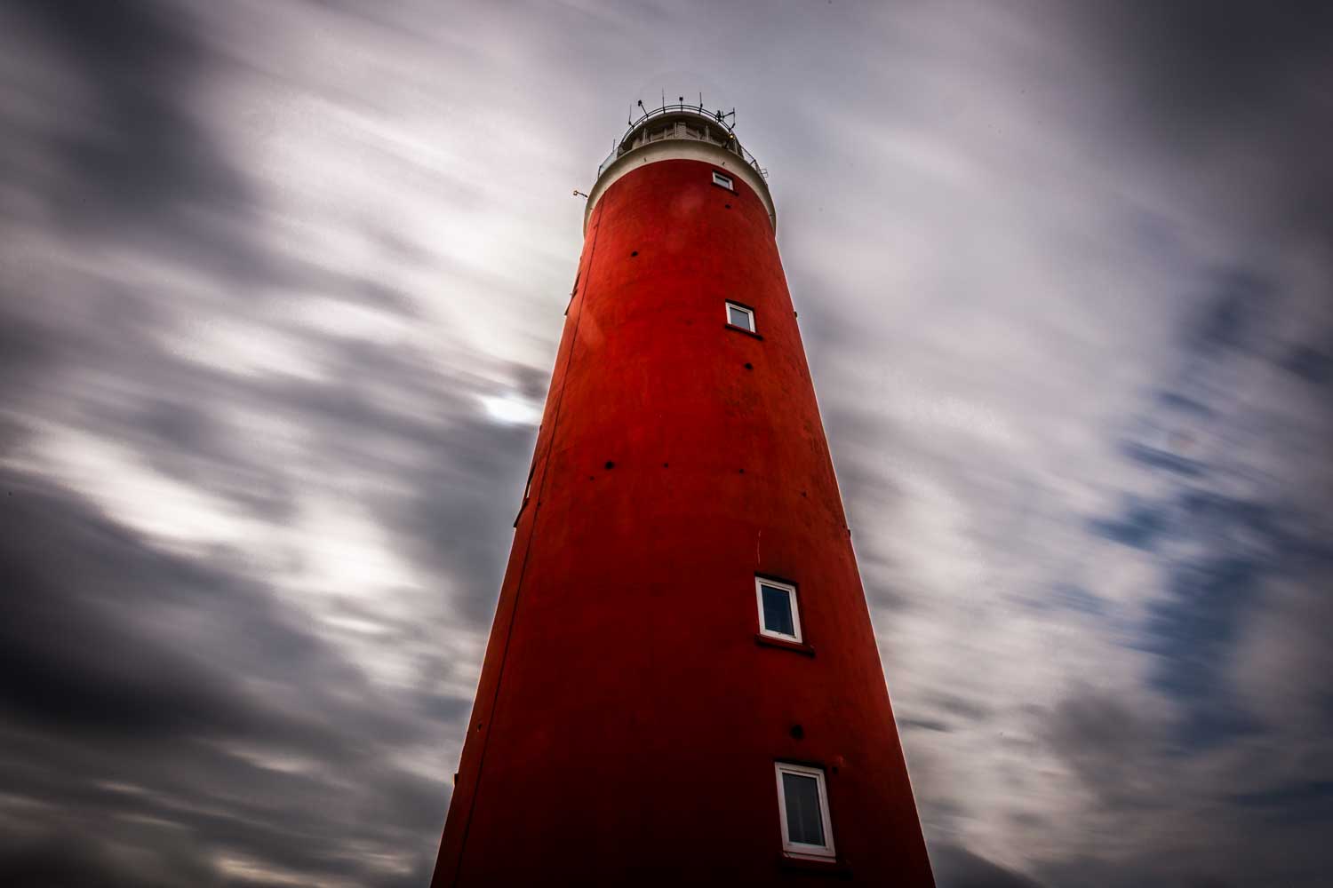 Tall red lighthouse tower against a cloudy sky, viewed from below, emphasizing its height and dramatic weather.