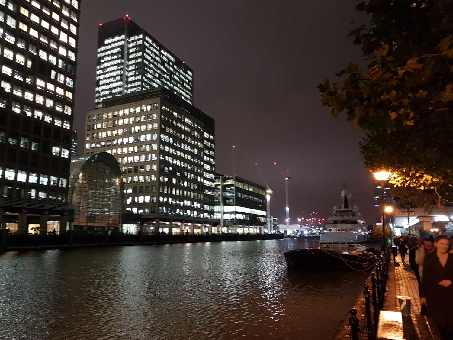 Night view of illuminated skyscrapers and a docked yacht on a riverside promenade, with people walking along the path.