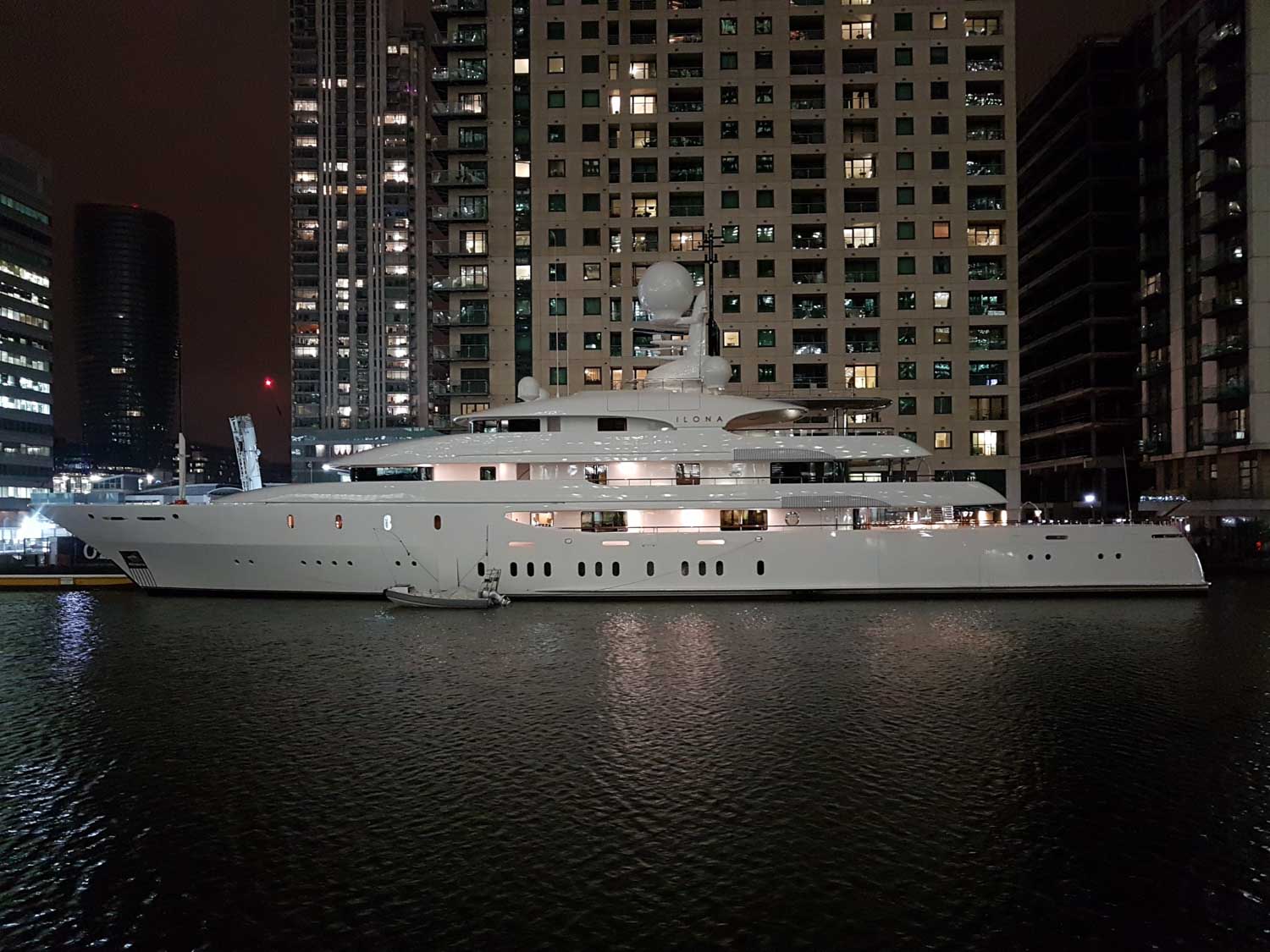 Luxurious yacht docked at night in an urban marina, surrounded by illuminated high-rise buildings.