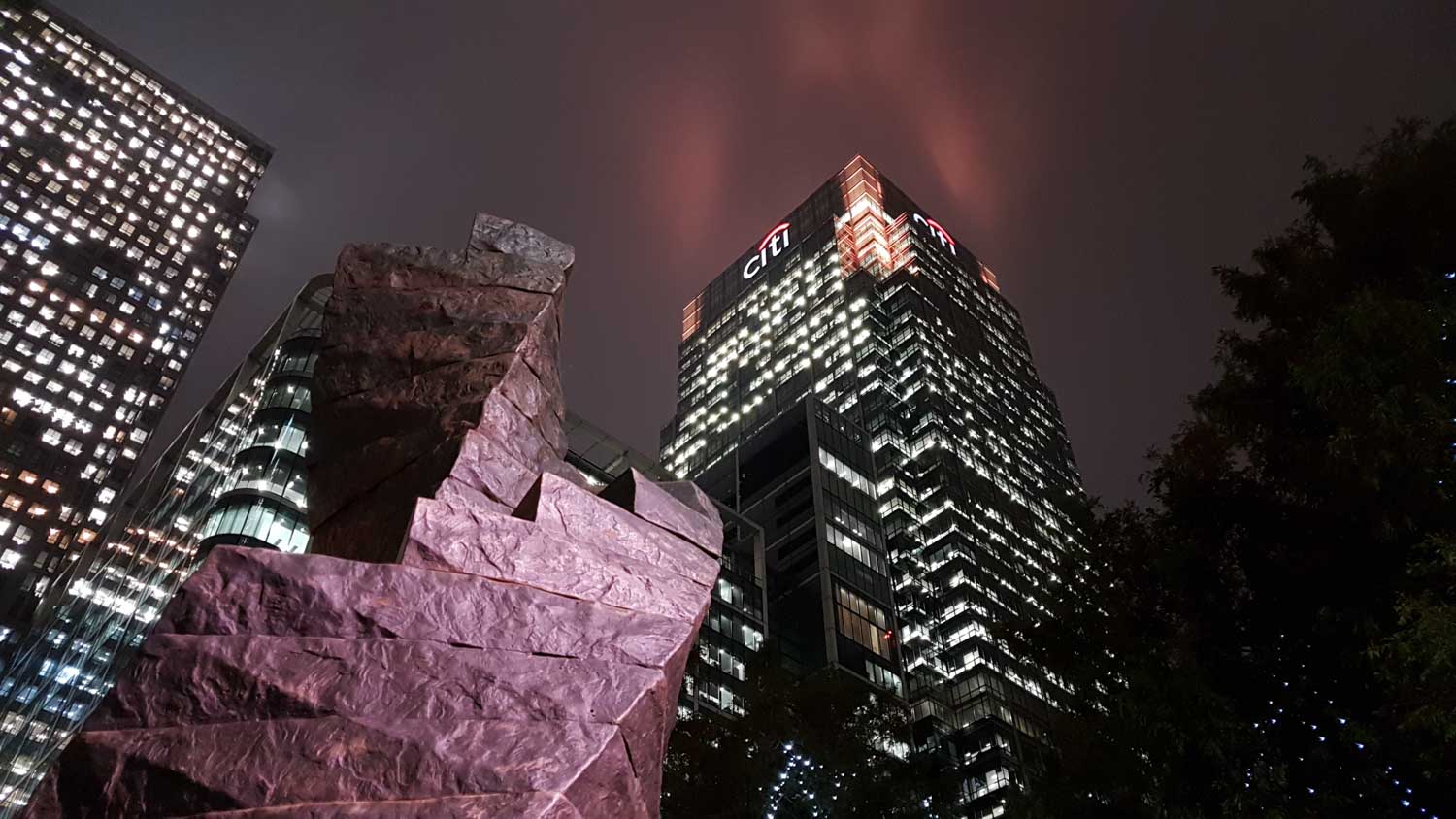 Modern skyscrapers lit at night with visible Citi logo, a large stone structure in the foreground, and a cloudy sky.