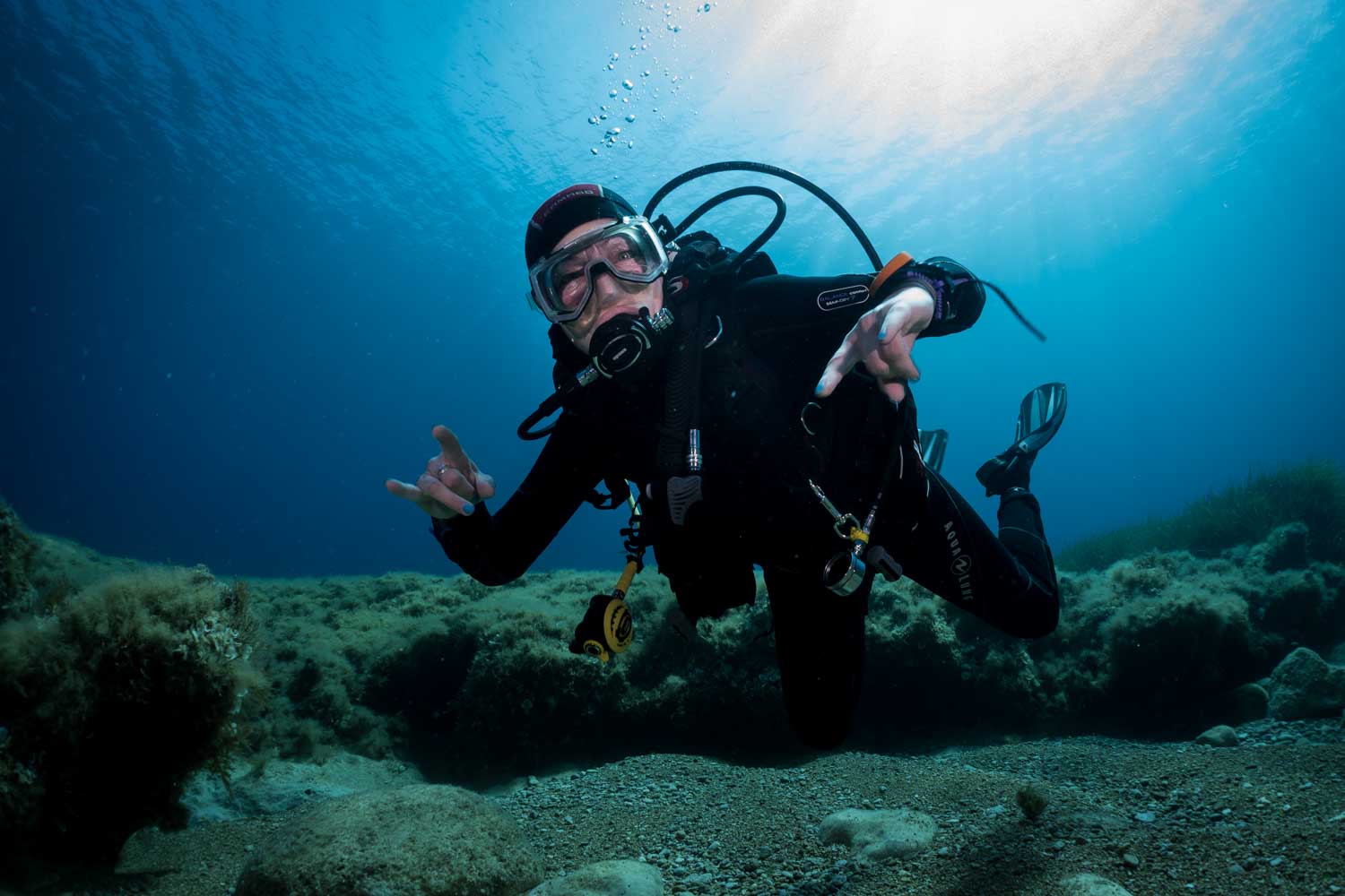 Scuba diver in black gear underwater gives hand signals above rocky ocean floor with sunlight filtering through.