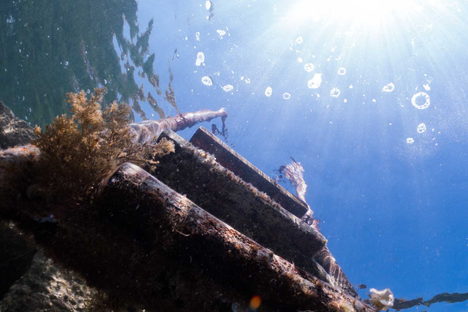 Underwater view of rusted shipwreck with sunlight streaming through water, highlighting coral growth and bubbles.