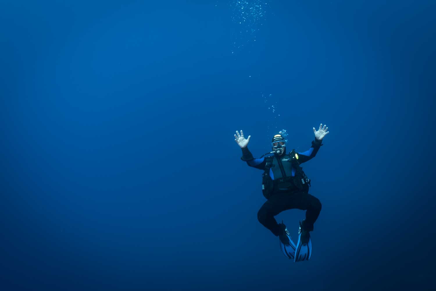 Scuba diver in blue gear underwater, with arms raised and bubbles rising above in deep blue ocean.