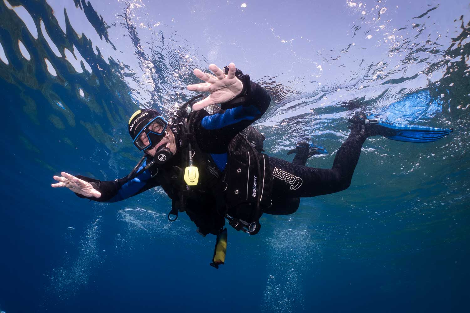 Scuba diver in blue ocean, wearing full gear, smiling and waving under clear water.