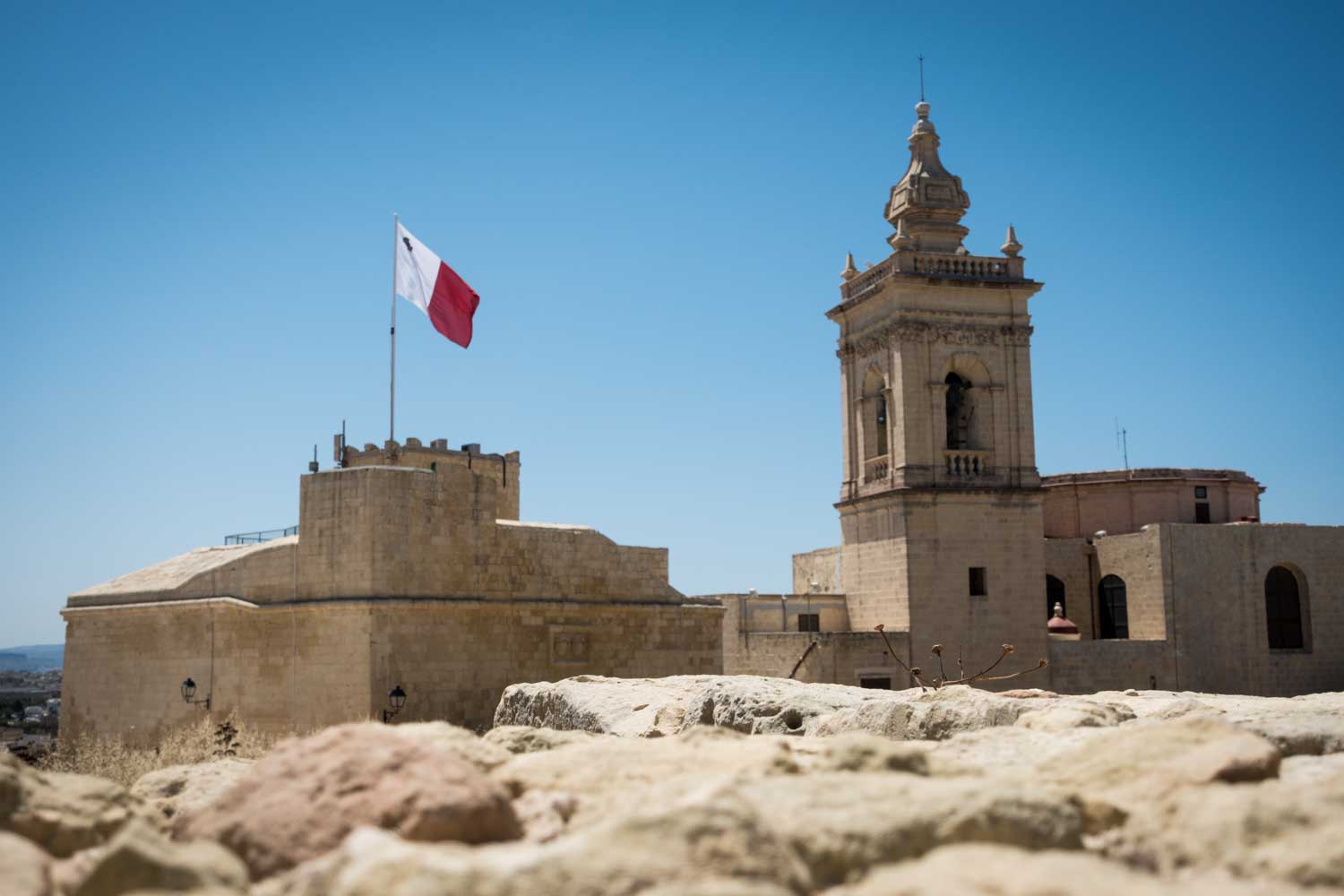 Historic stone tower with a Maltese flag under clear blue skies, surrounded by ancient fortifications.