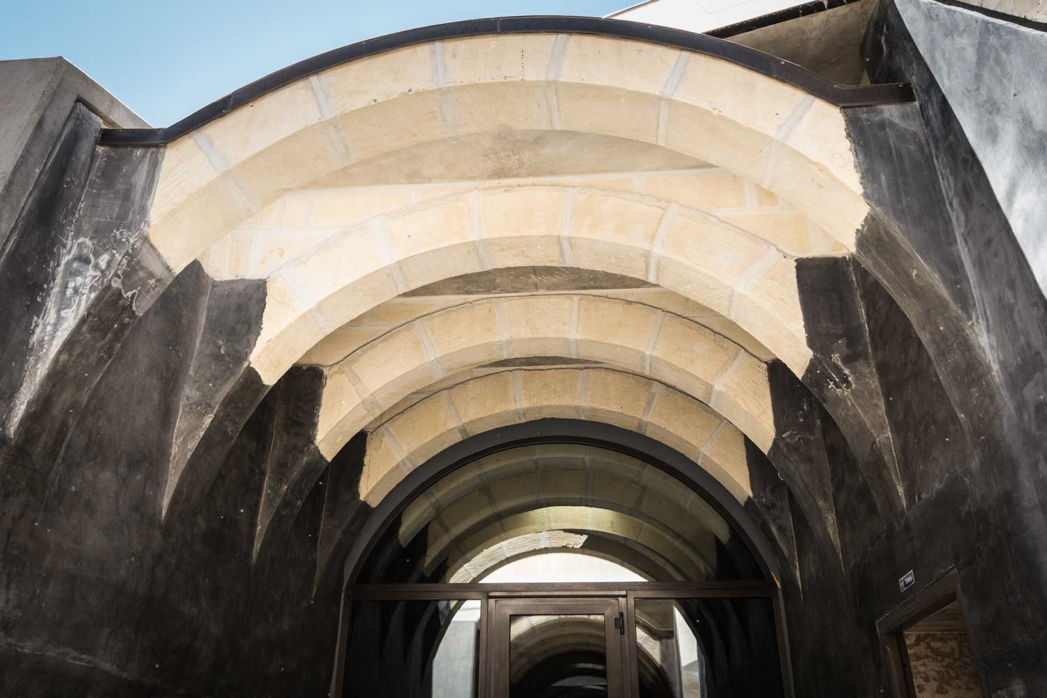 Modern arched entrance with layered stone and dark walls under a clear blue sky