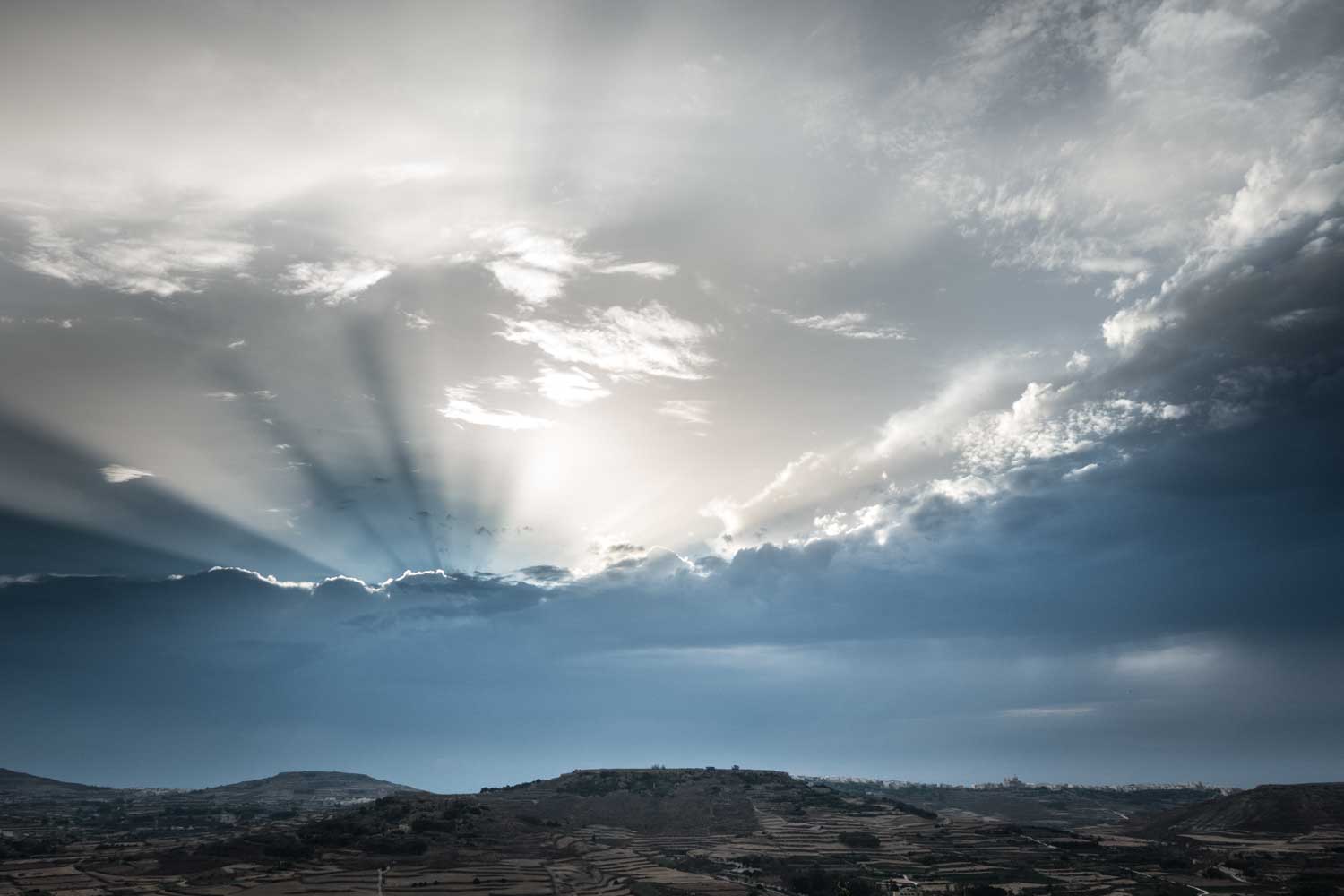 Dramatic sun rays break through clouds over a hilly landscape at sunset.