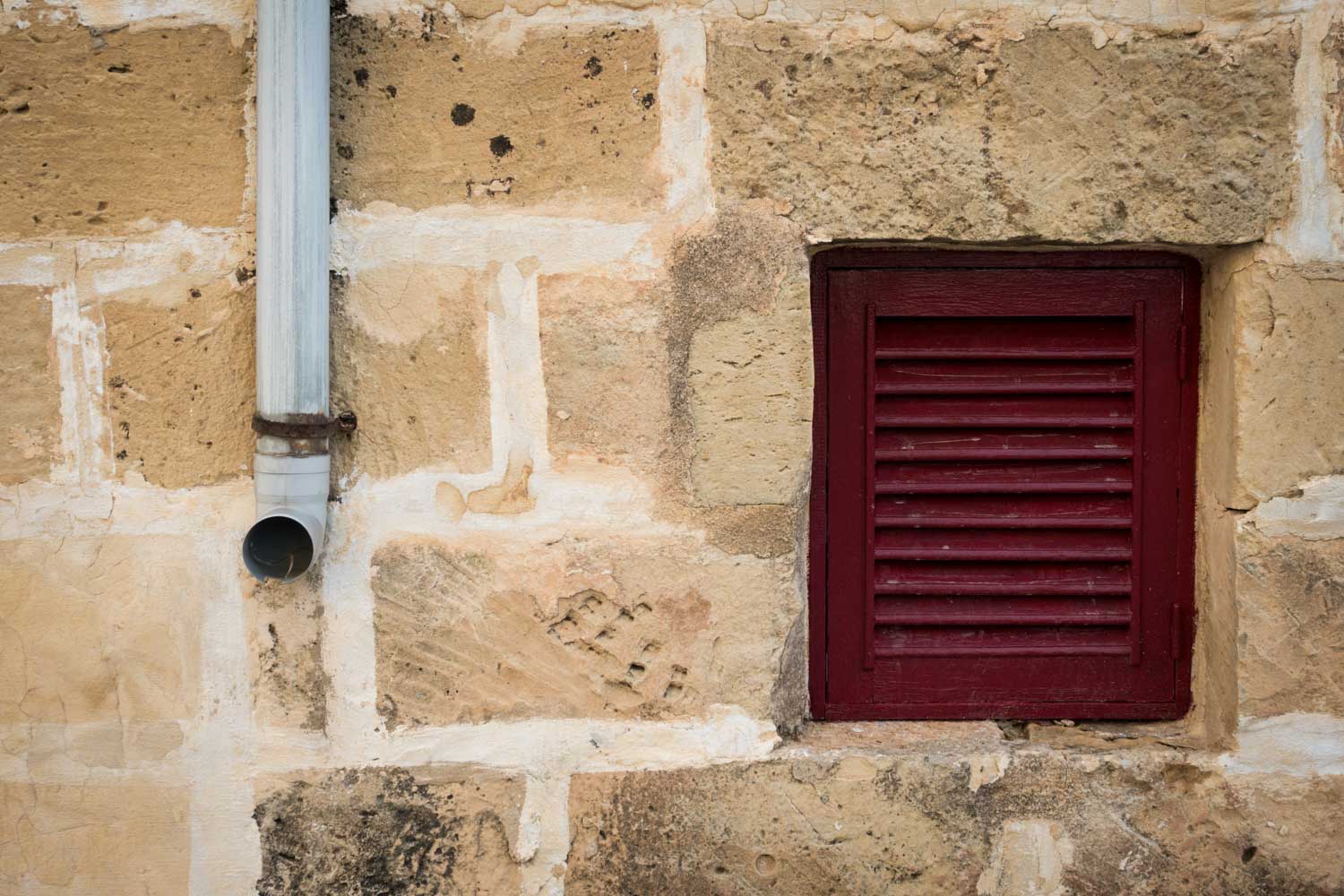 Stone wall with a red shuttered window and adjacent gray downspout.