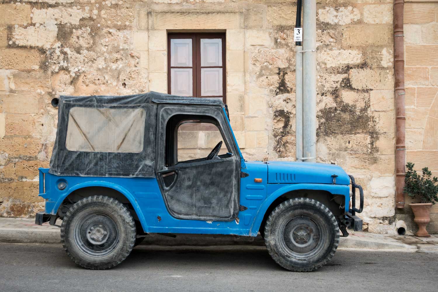 Rustic blue jeep with black soft top parked by an old stone wall on a quiet street.