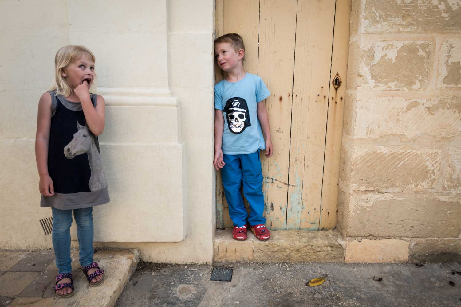 Two children standing against a rustic wall; a girl in a horse-print dress and a boy in a skull t-shirt smiling at each other.