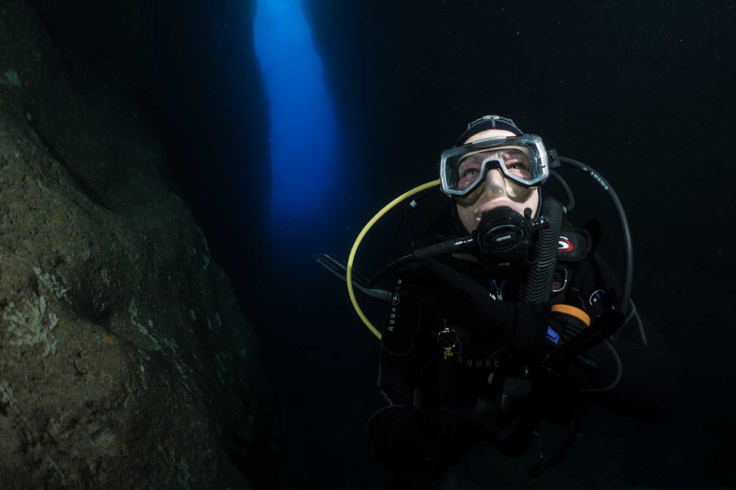 Scuba diver explores deep underwater cave, illuminated by a shaft of blue light.
