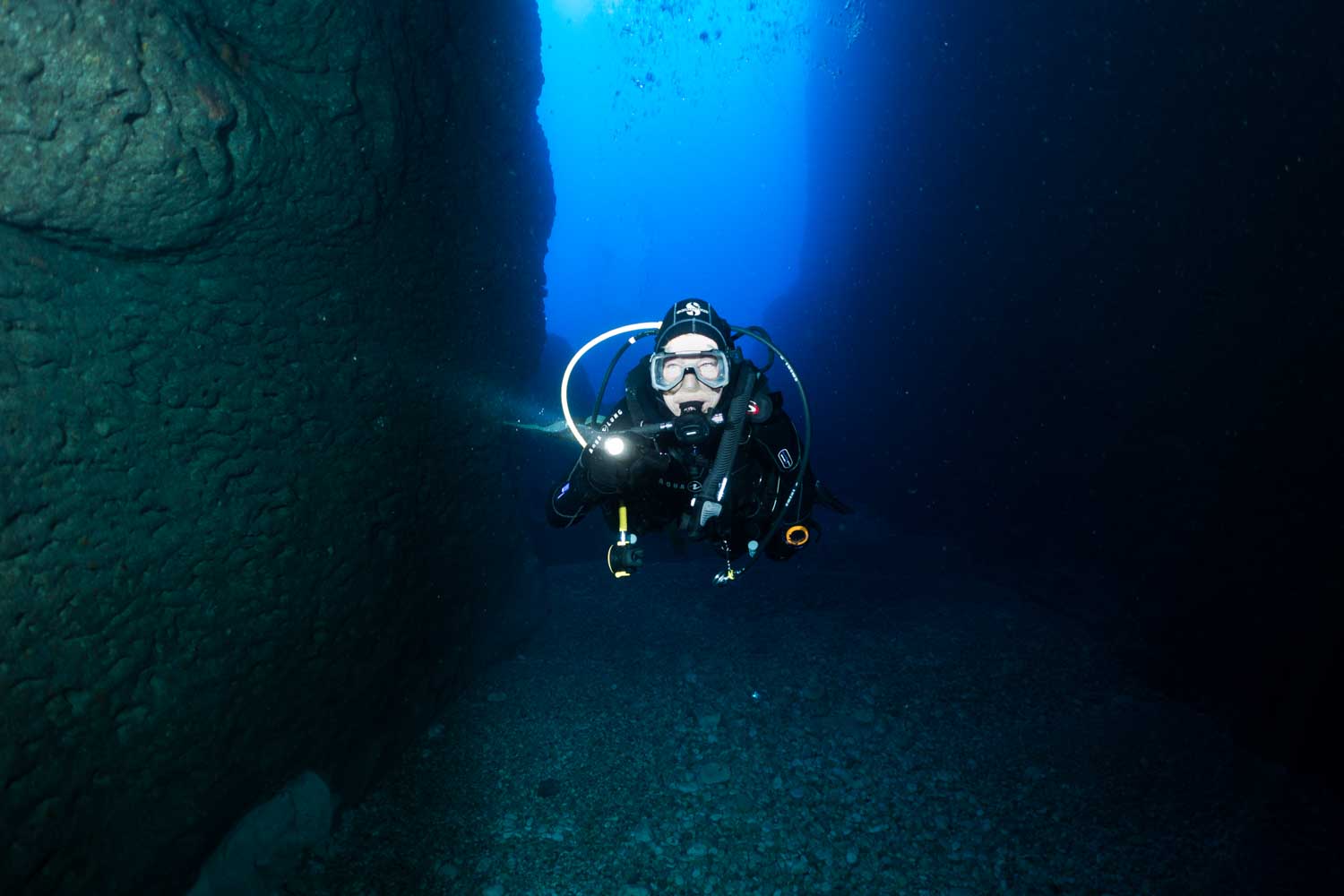 Scuba diver explores underwater canyon with flashlight, surrounded by deep blue water and rocky walls.
