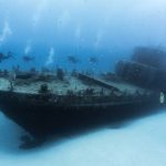 Divers exploring an underwater shipwreck surrounded by fish in clear blue ocean water.