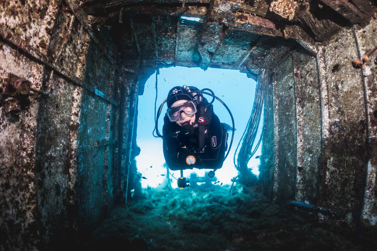 Scuba diver exploring underwater shipwreck interior with clear blue water and marine growth.