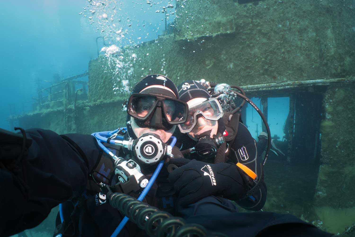 Two scuba divers take a selfie underwater near a sunken shipwreck, wearing diving gear and goggles.