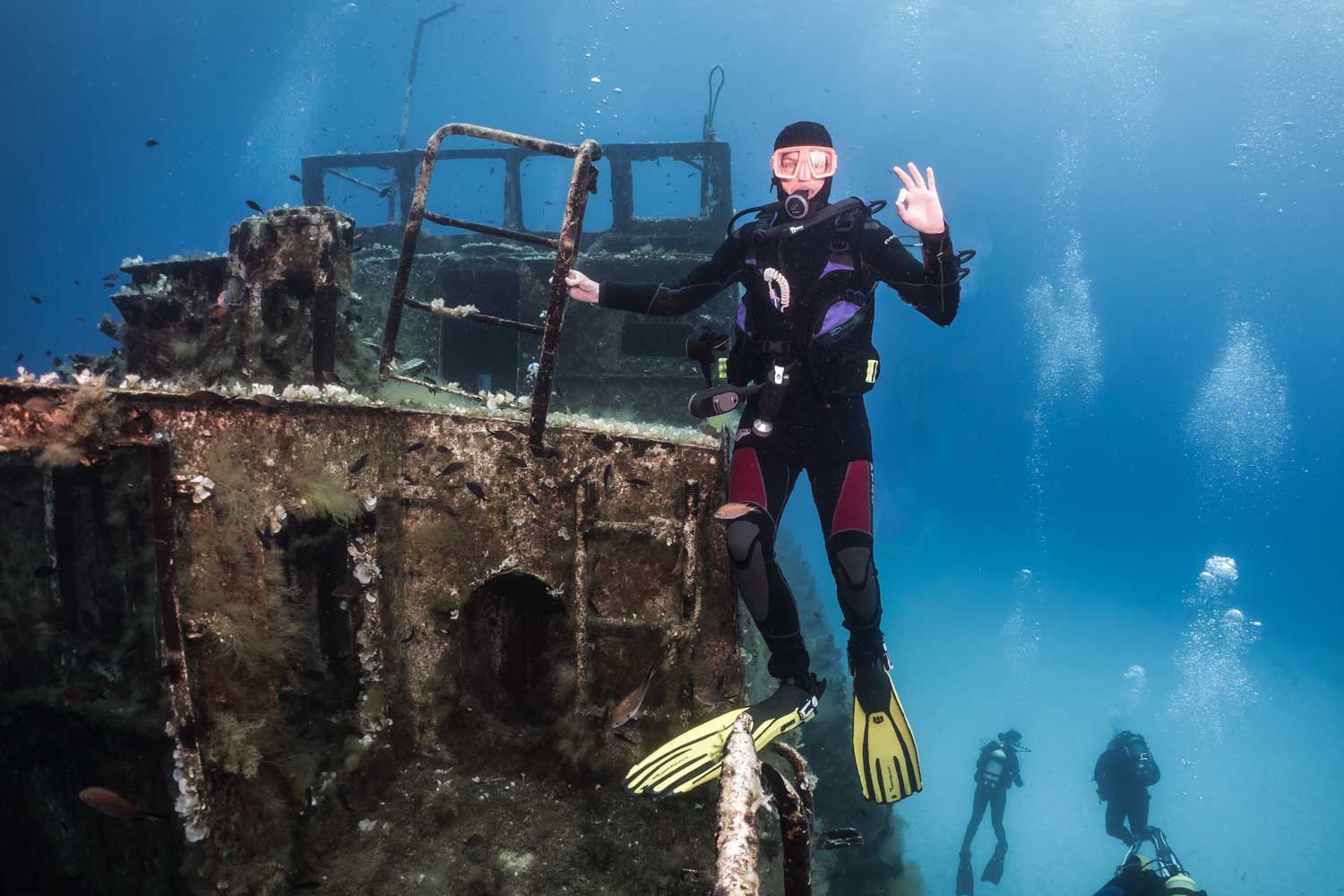 Diver exploring a sunken shipwreck underwater, surrounded by bubbles and small fish, signaling OK with hand gesture.
