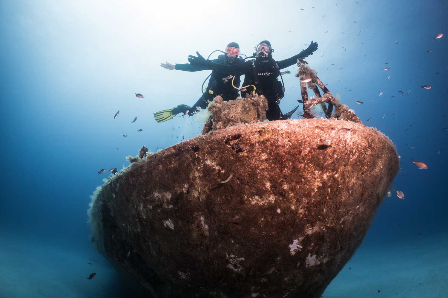 Scuba divers explore a sunken shipwreck surrounded by fish in clear blue ocean water.
