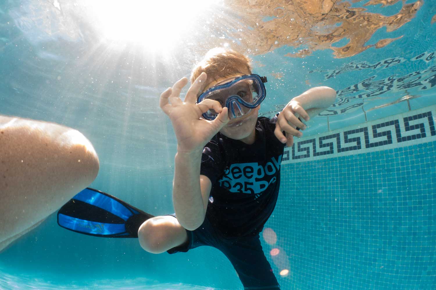 Child underwater in a pool, wearing goggles and snorkel gear, gives an OK sign with hand. Sunlight filters through water.