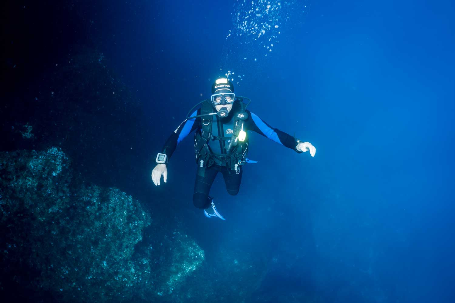 Scuba diver in deep blue ocean, wearing diving gear and surrounded by underwater rock formations.