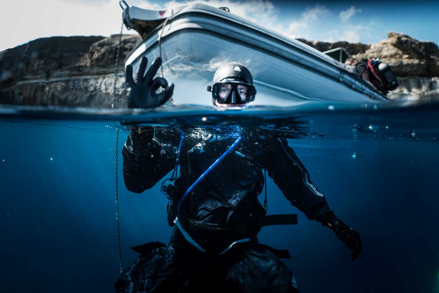 Scuba diver underwater, giving an OK sign near a boat, with rocky coastline in the background.