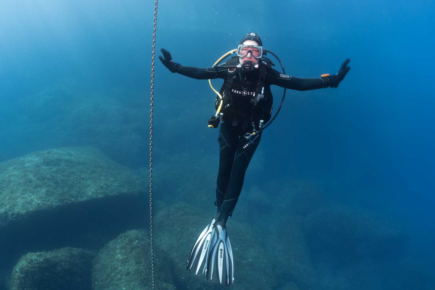 Scuba diver with arms open underwater near rocks and chain, wearing gear and fins, in clear blue ocean.