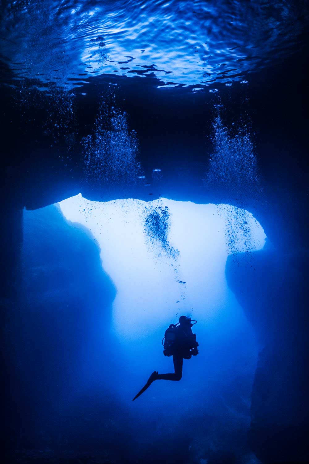 Scuba diver exploring underwater cave with bubbles rising to the surface, silhouetted against deep blue water.