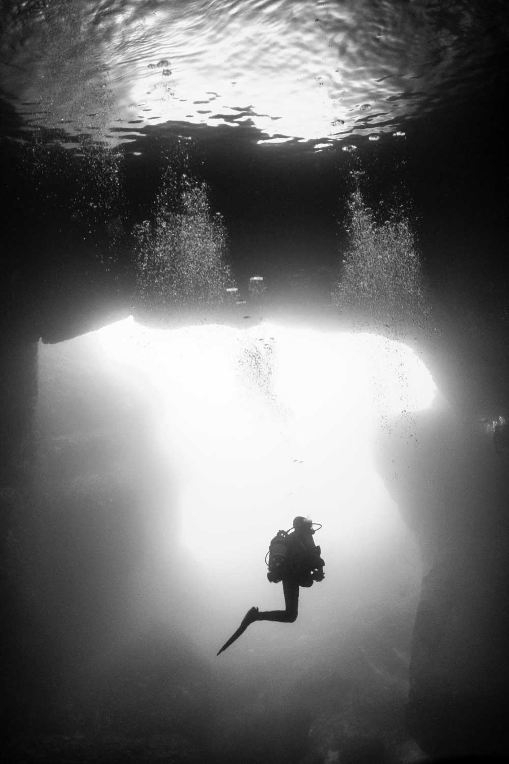 Scuba diver exploring underwater cave with air bubbles rising, silhouetted against bright light from cave opening.