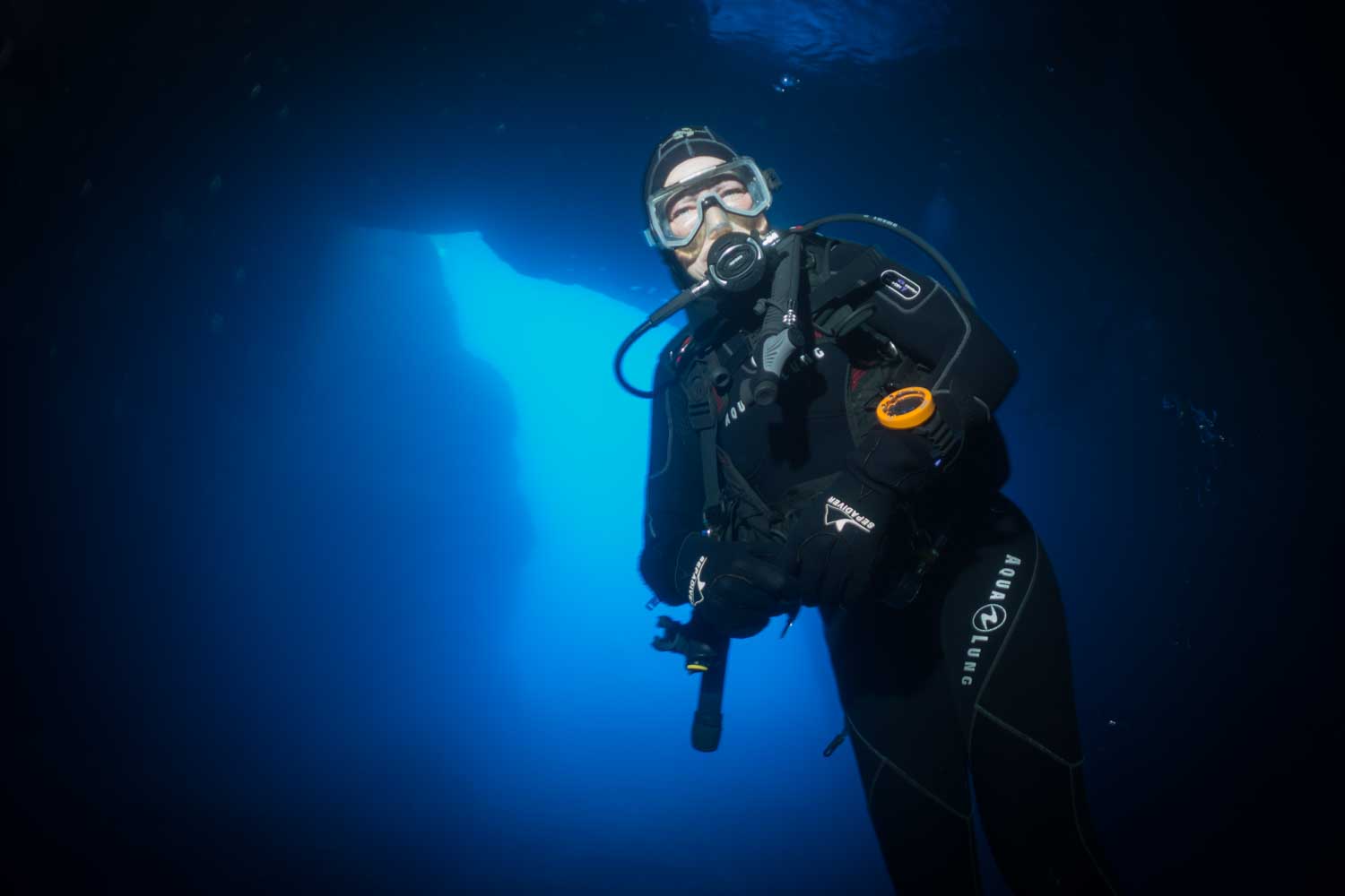 Scuba diver in black wetsuit explores underwater cave, illuminated by deep blue light.