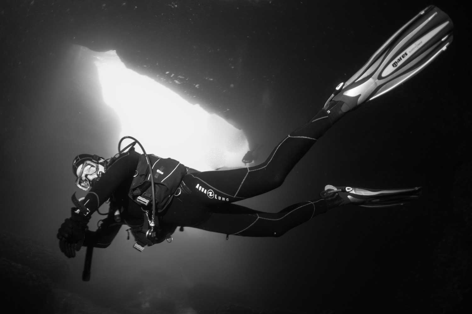 Scuba diver explores underwater cave in wetsuit and fins, illuminated entrance visible above in black and white.