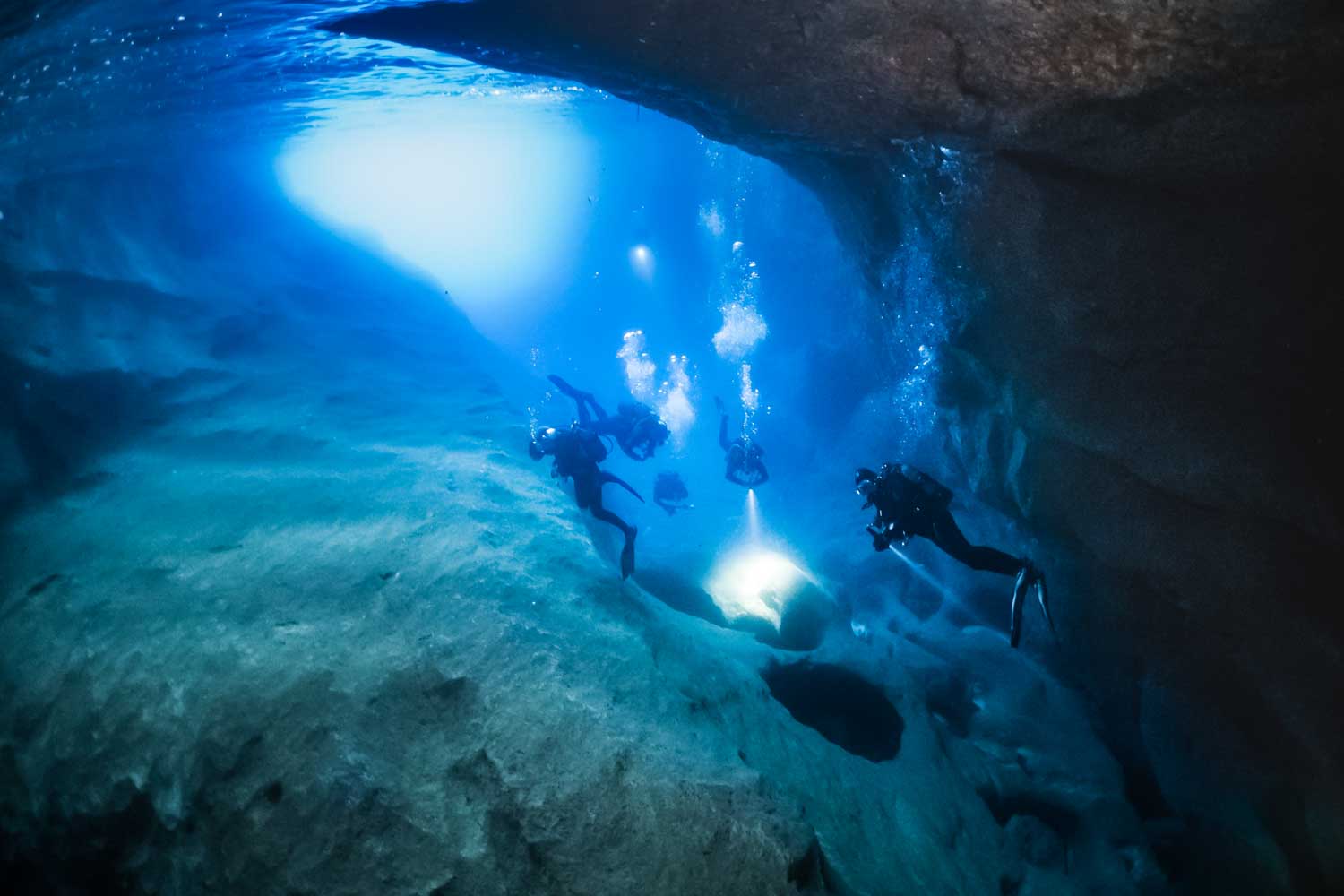 Divers exploring an underwater cave with flashlights illuminating the blue water around them.