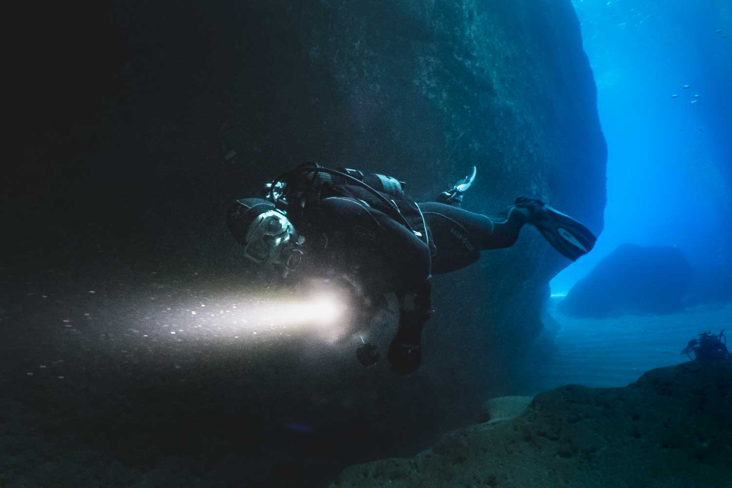 Scuba diver explores underwater cave with flashlight in deep blue ocean.