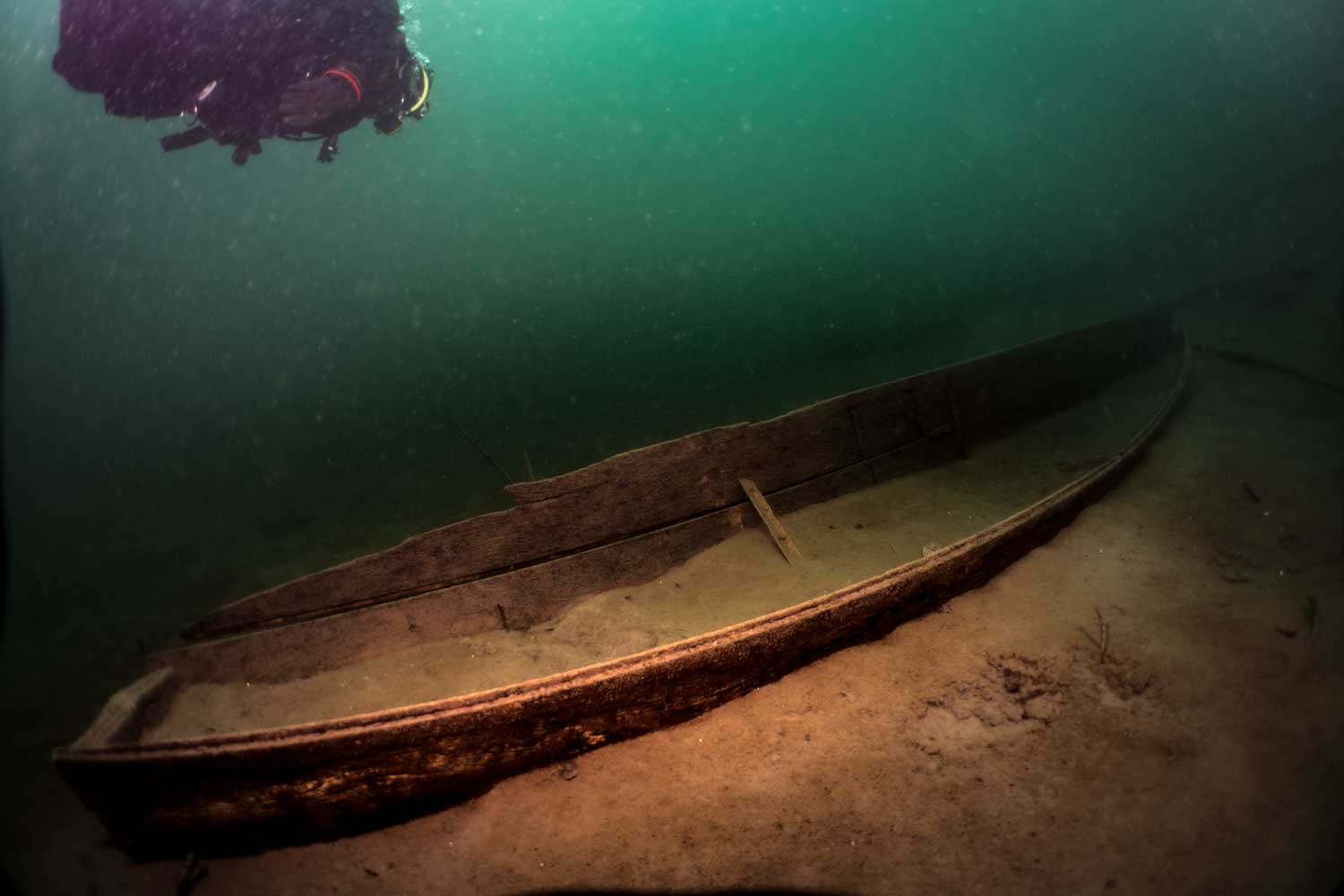 Scuba diver explores submerged wooden shipwreck on sandy ocean floor under greenish water.