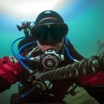 Scuba diver in red drysuit underwater, using a rebreather and capturing a selfie in greenish-blue water.