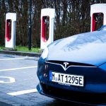 Blue Tesla Model S parked at a charging station with four Tesla Supercharger units in the background.
