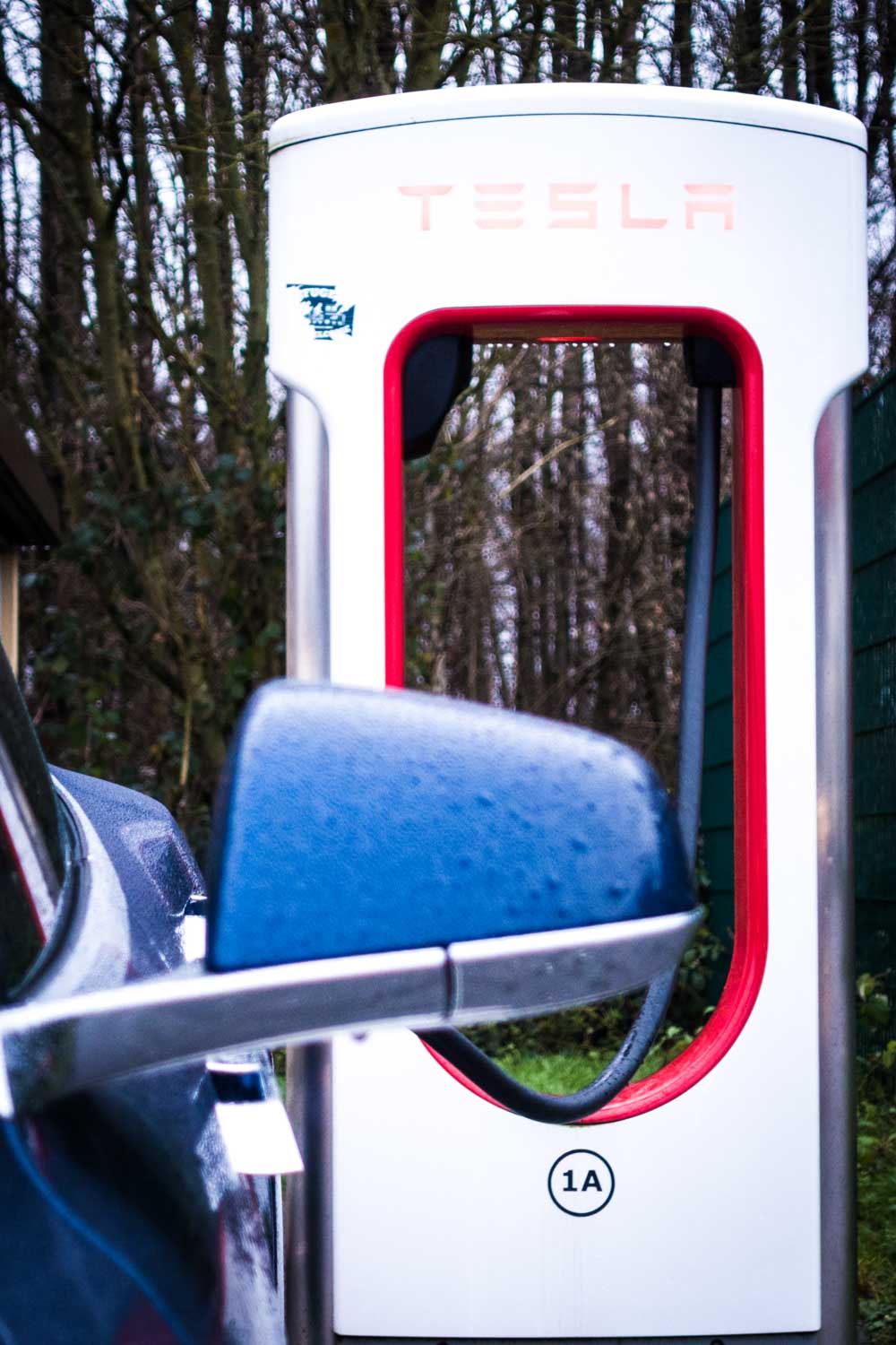 Tesla car at a charging station with trees in the background, showing a close-up of a side mirror.